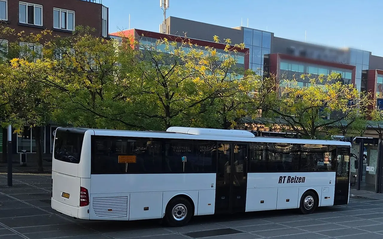 Bus van RT Reizen op een busplein bij een station, in de avondzon, met bomen en kantoren op de achtergrond.