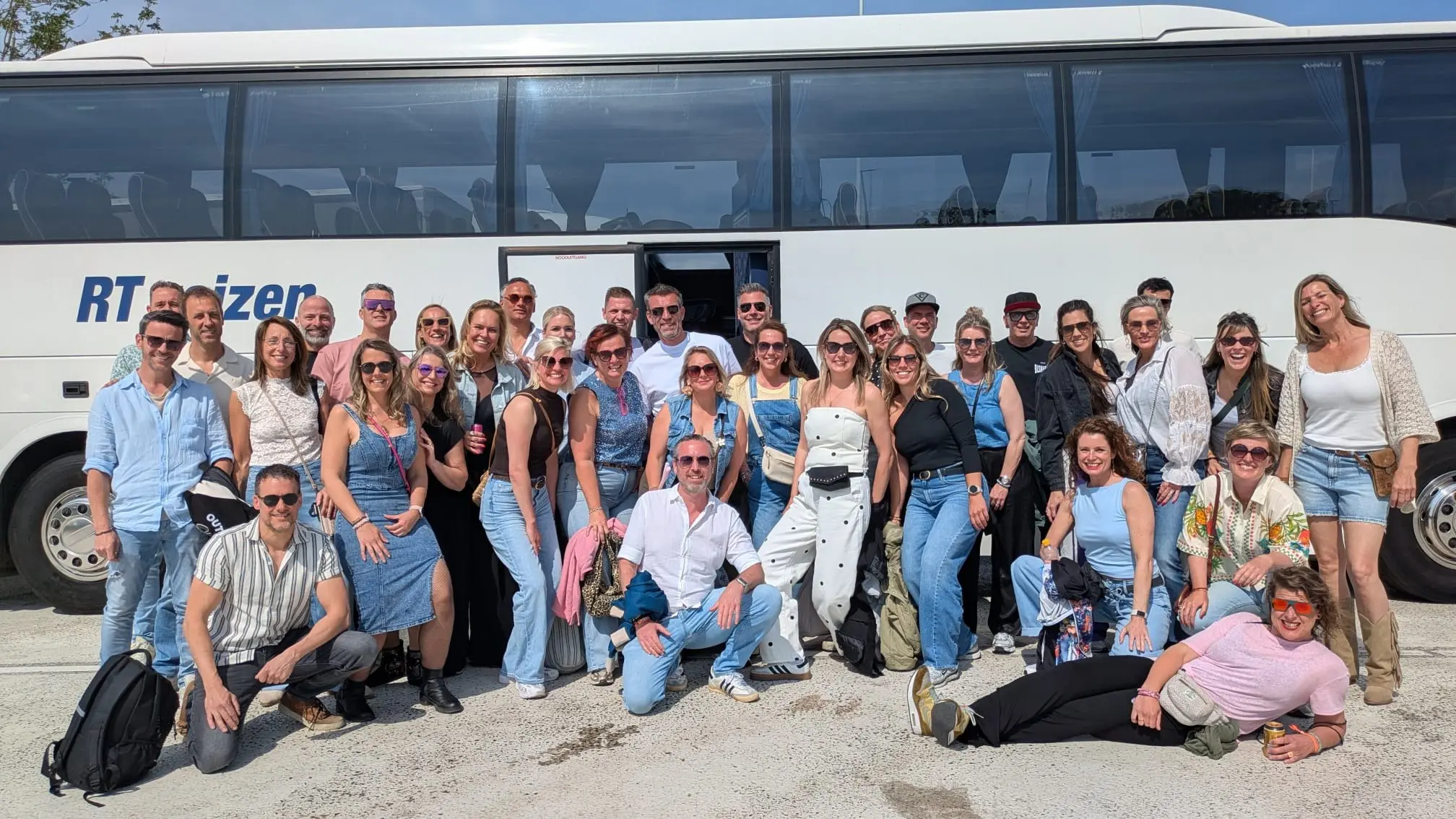 Een groep gasten poseert lachend voor de touringcar van RT Reizen bij het begin van hun uitje.