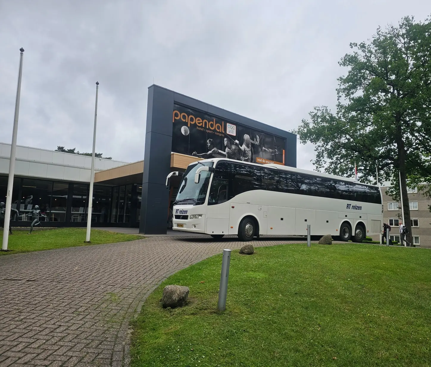 De witte touringcar van RT Reizen staat voor de ingang van sportcentrum Papendal, onder het bord met sporters.