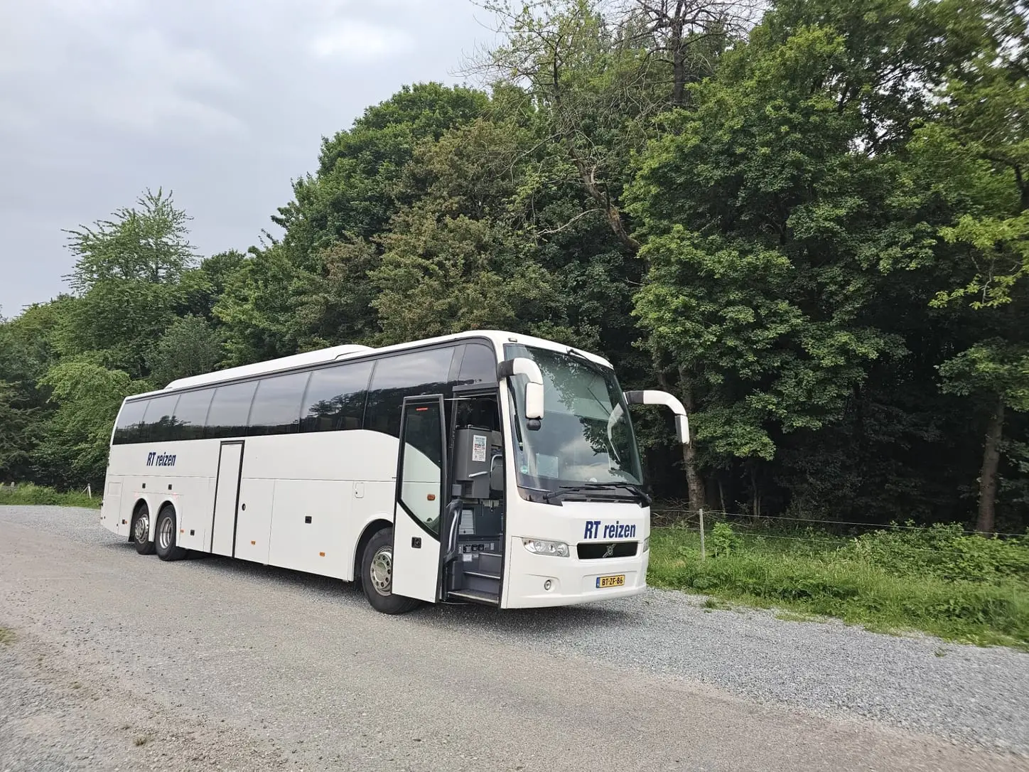 De touringcar van RT Reizen staat met de deur open op een landweg langs een bos, klaar om de groep te laten instappen.
